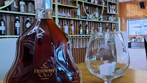 Una botella de coñac Hennessy y una copa sobre una mesa de madera en un bar, con estantes llenos de botellas al fondo.