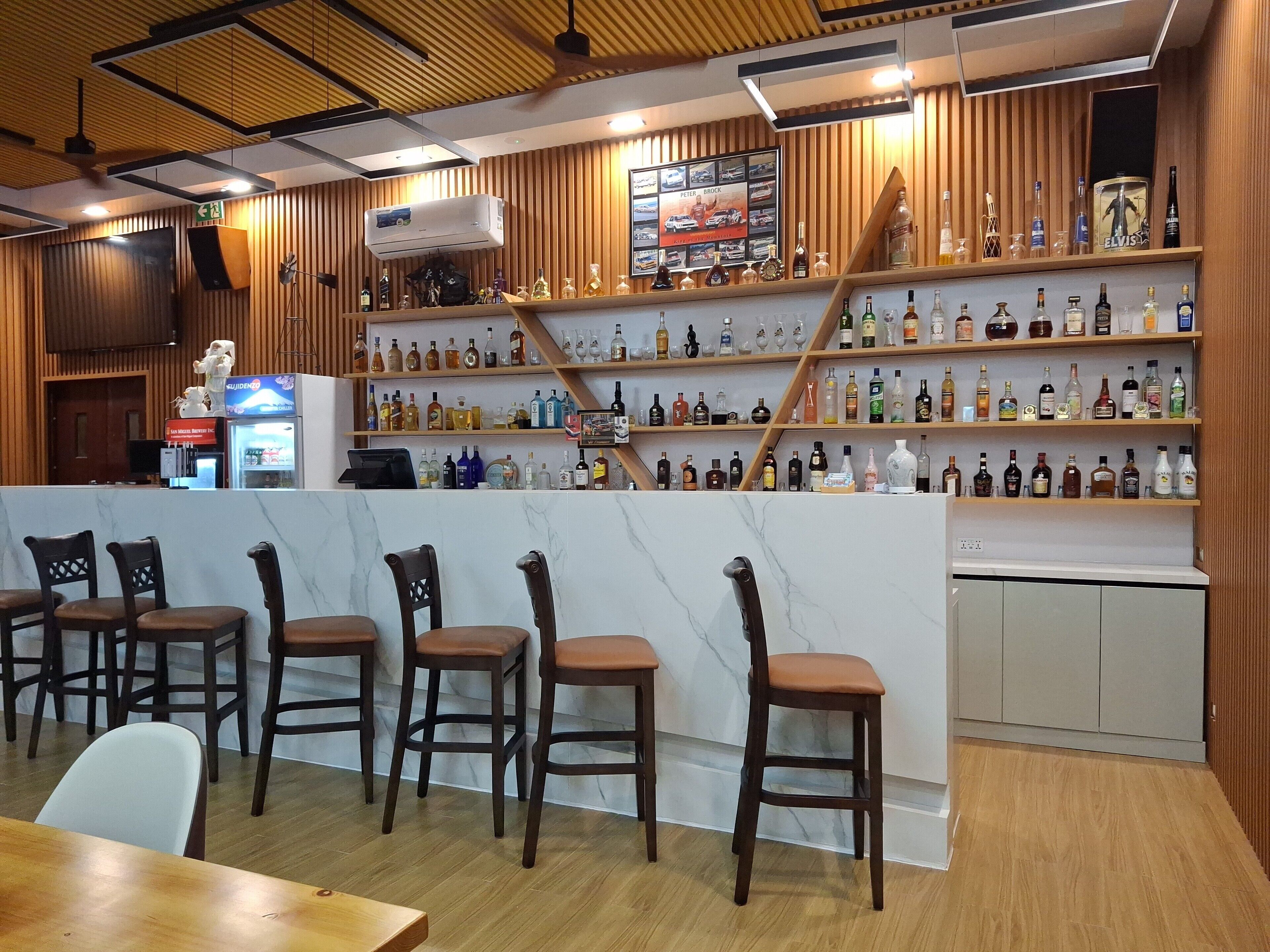 A bar area with wooden shelves stocked with various liquor bottles, a marble counter, and several wooden bar stools.
