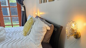 Un lit bien fait, avec une couette blanche, un oreiller marron et un oreiller décoratif jaune. On y trouve une table de chevet avec un vase de fleurs et une bougie. La pièce est dotée d’une fenêtre avec des rideaux, d’une lampe fixée au mur et de trois œuvres d’art encadrées.