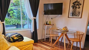 Un salon confortable avec un canapé, une table à manger et des chaises, un téléviseur fixé au mur et une fenêtre avec des rideaux.