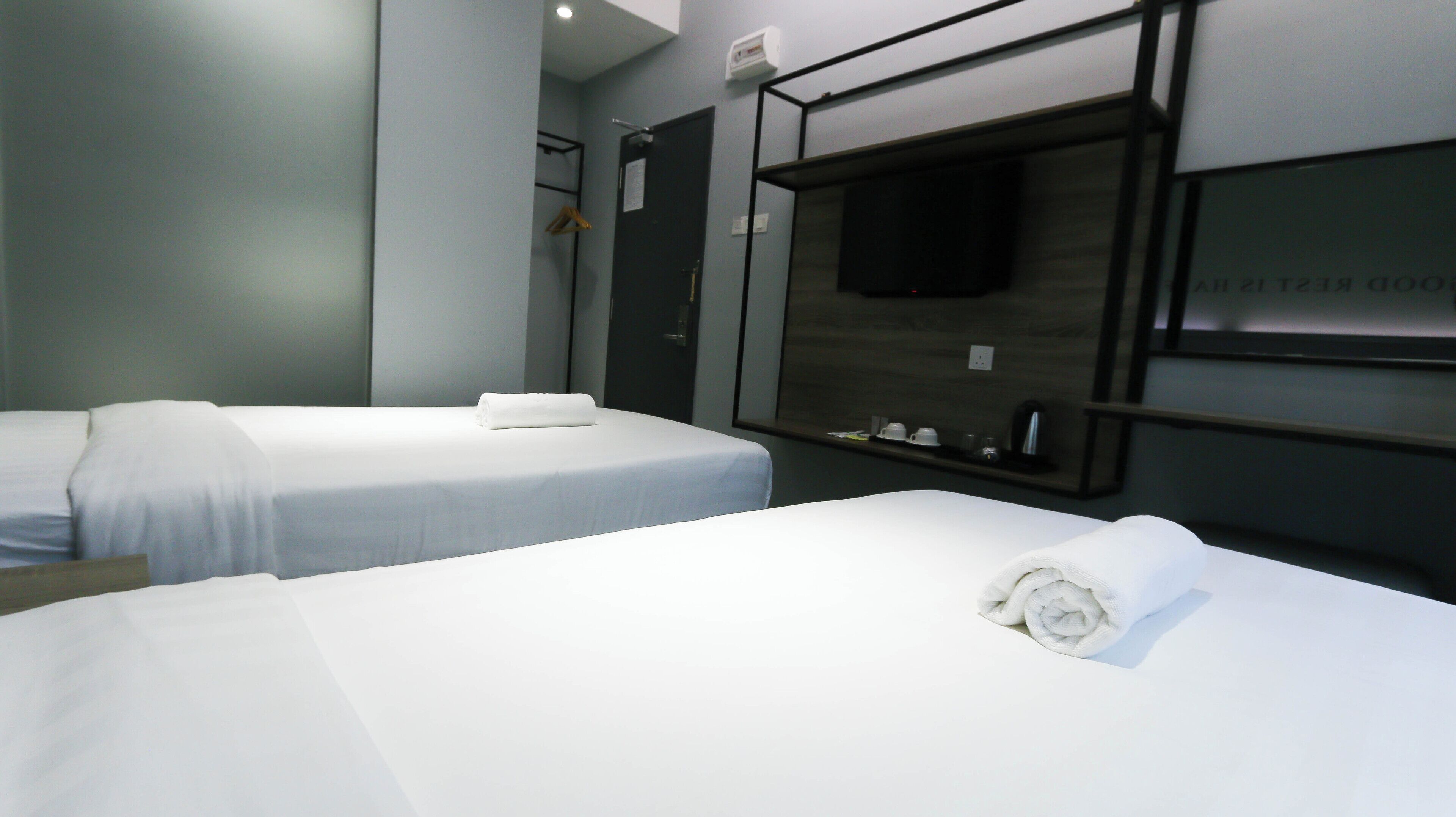 A hotel room with two single beds, a TV mounted on the wall, a small shelf with toiletries, and a door with a towel hanging on it.