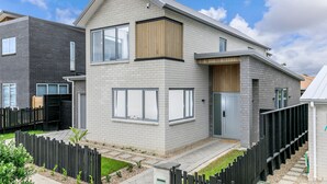 Une maison moderne à deux étages, dotée d’un toit plat, de grandes fenêtres et d’un petit jardin à l’avant, délimité par une clôture en bois.