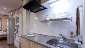 Une cuisine moderne équipée d’un évier en acier inoxydable, d’un four à micro-ondes et d’un réfrigérateur. On aperçoit une fenêtre avec des stores et un lit au fond de la pièce.