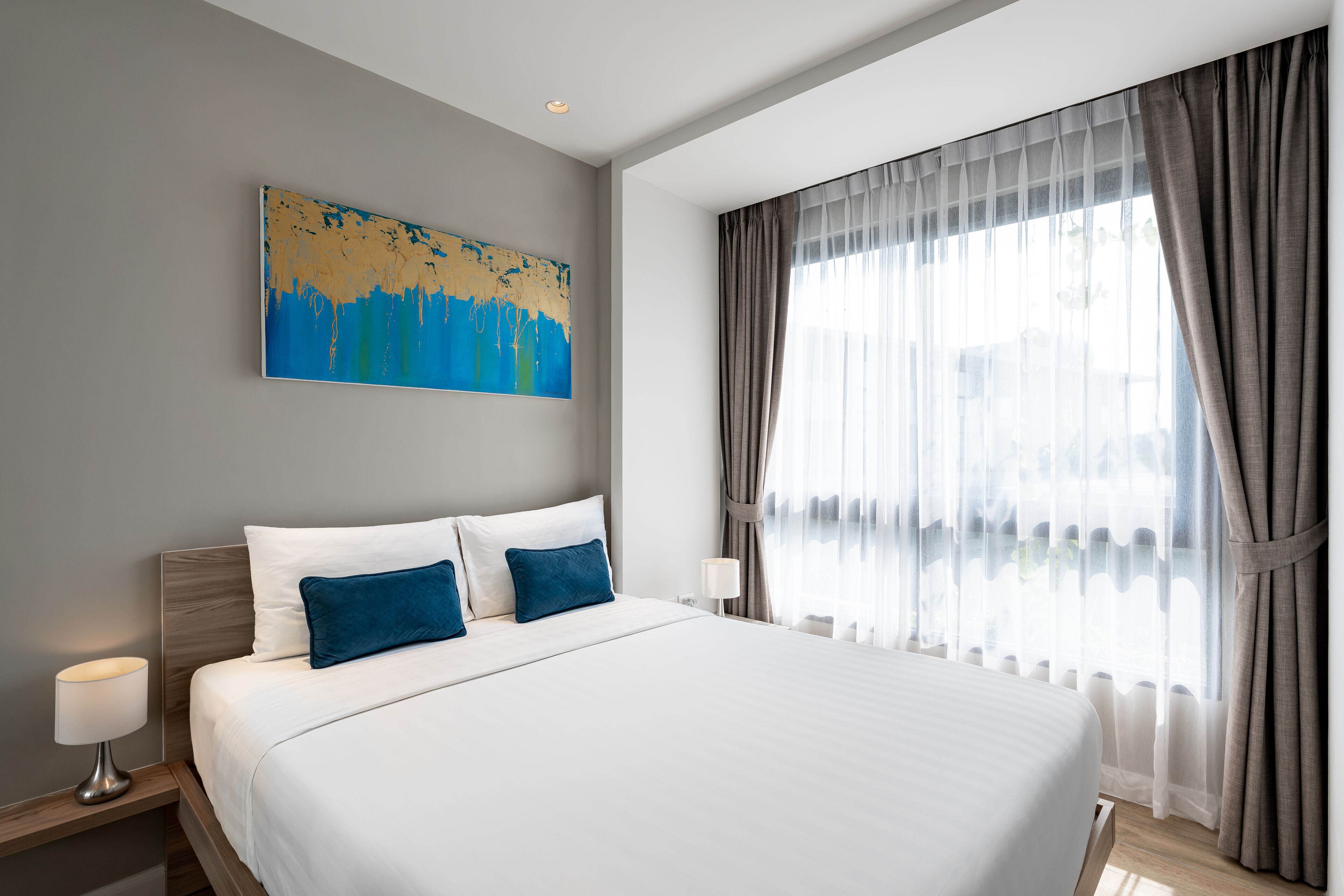 Habitación de hotel con una cama grande, una mesita de noche, una lámpara, un cuadro en la pared y una ventana con cortinas.