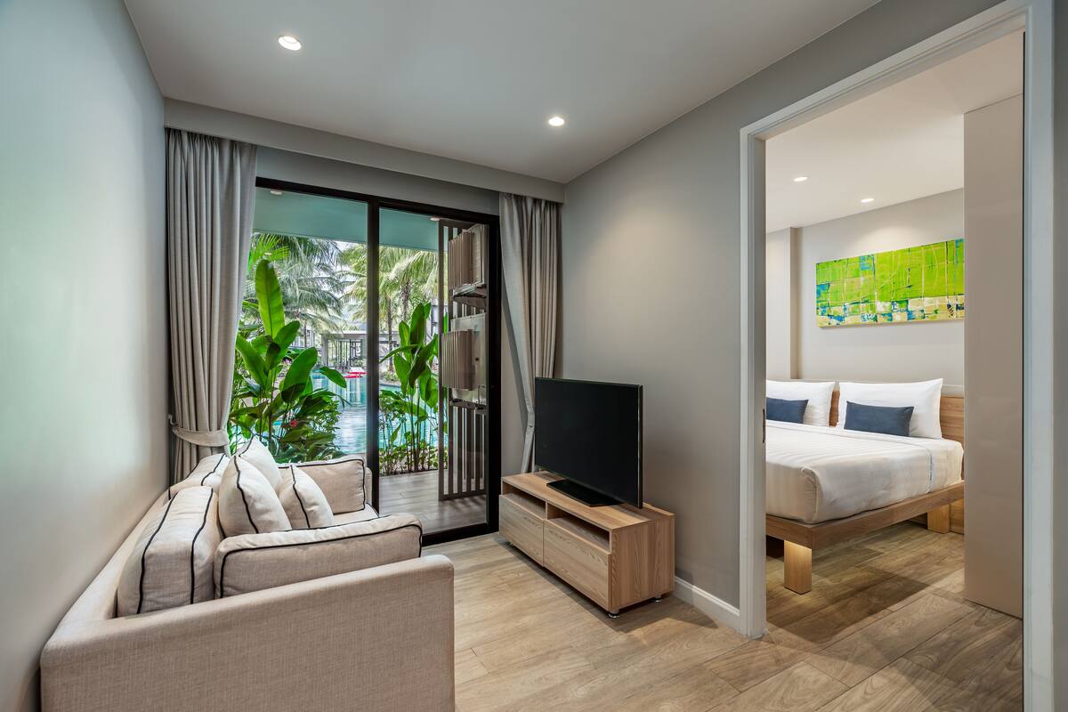 Quarto de hotel moderno com sofá, televisão em suporte de madeira, cama com roupa de cama branca e vista para palmeiras através de grandes janelas.