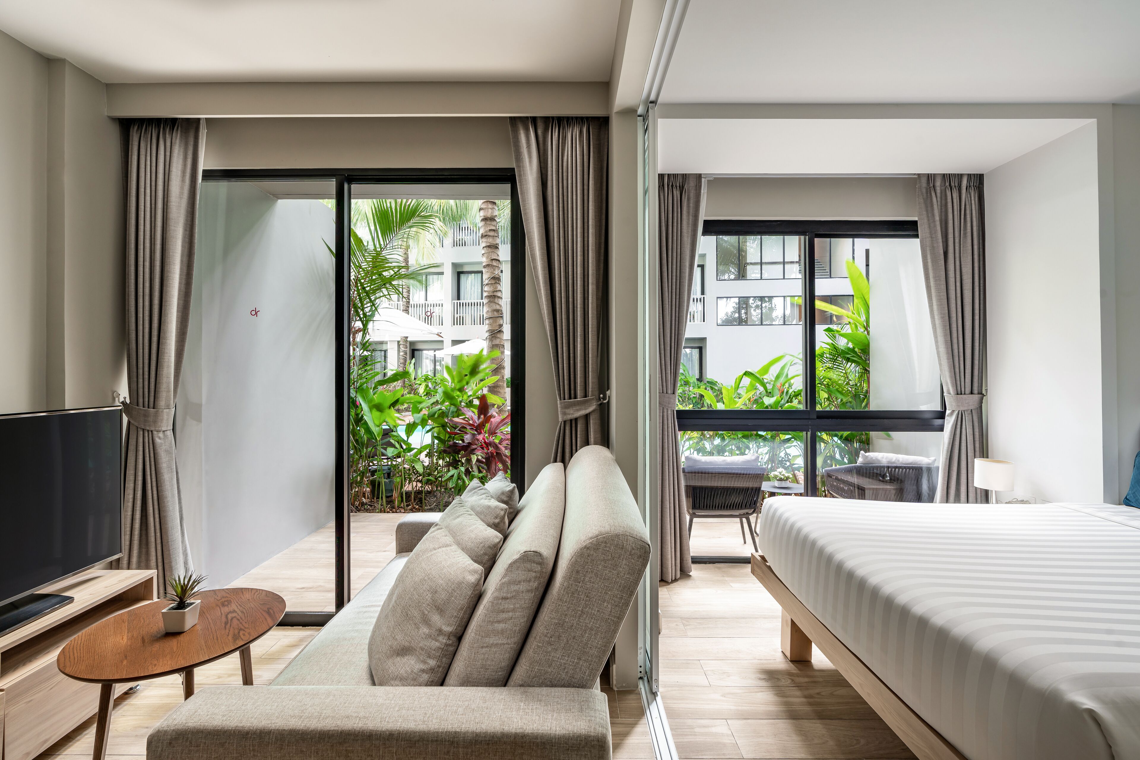 Ein modernes Hotelzimmer mit Sofa, Fernseher, einem kleinen Tisch mit Pflanze, einem Bett und einem Balkon mit Pflanzen und Blick auf Gebäude.