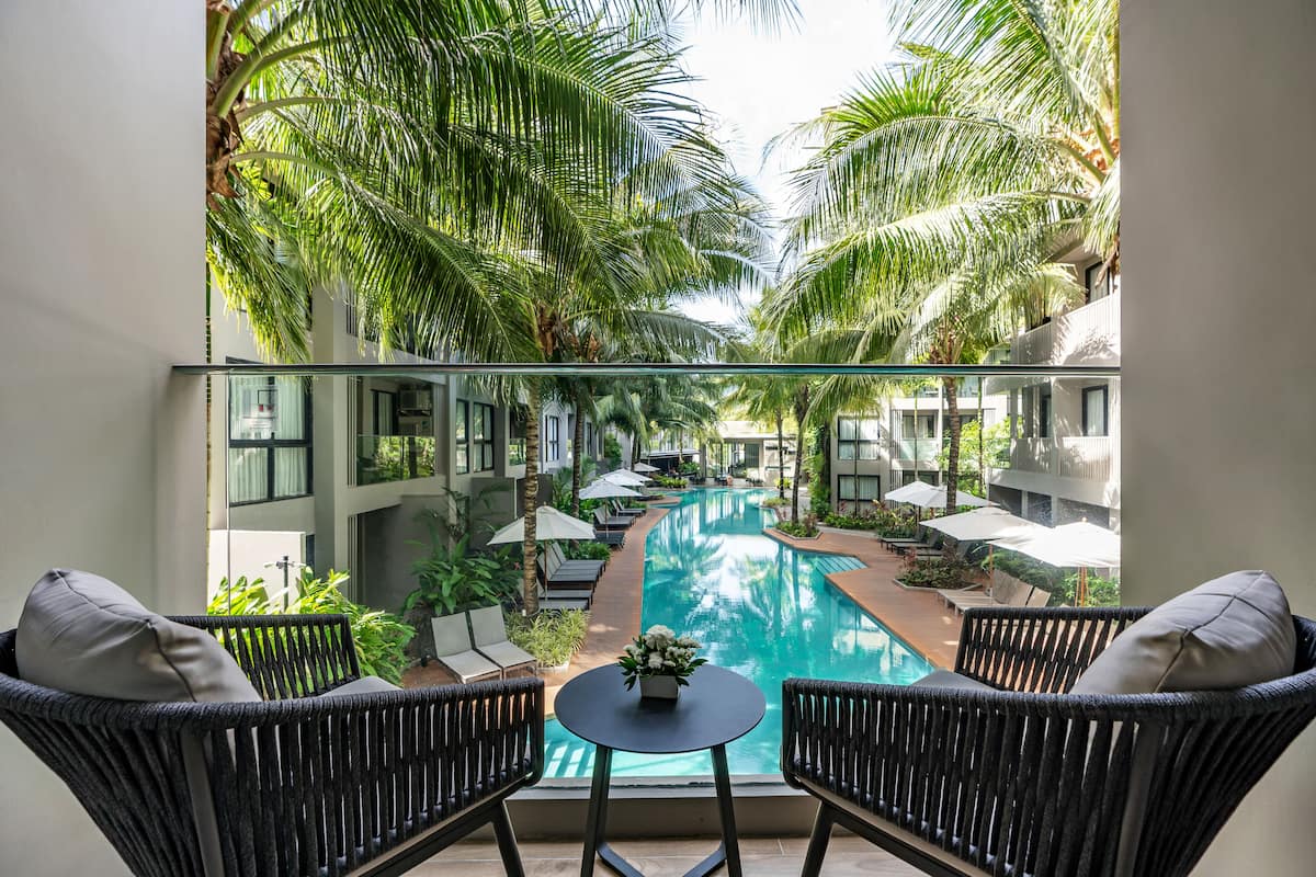 Uma varanda com cadeiras de vime e uma mesa pequena, com vista para uma piscina rodeada por palmeiras e espreguiçadeiras.