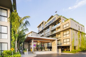 Hotel moderno com entrada de vidro, área de estar ao ar livre e vegetação.