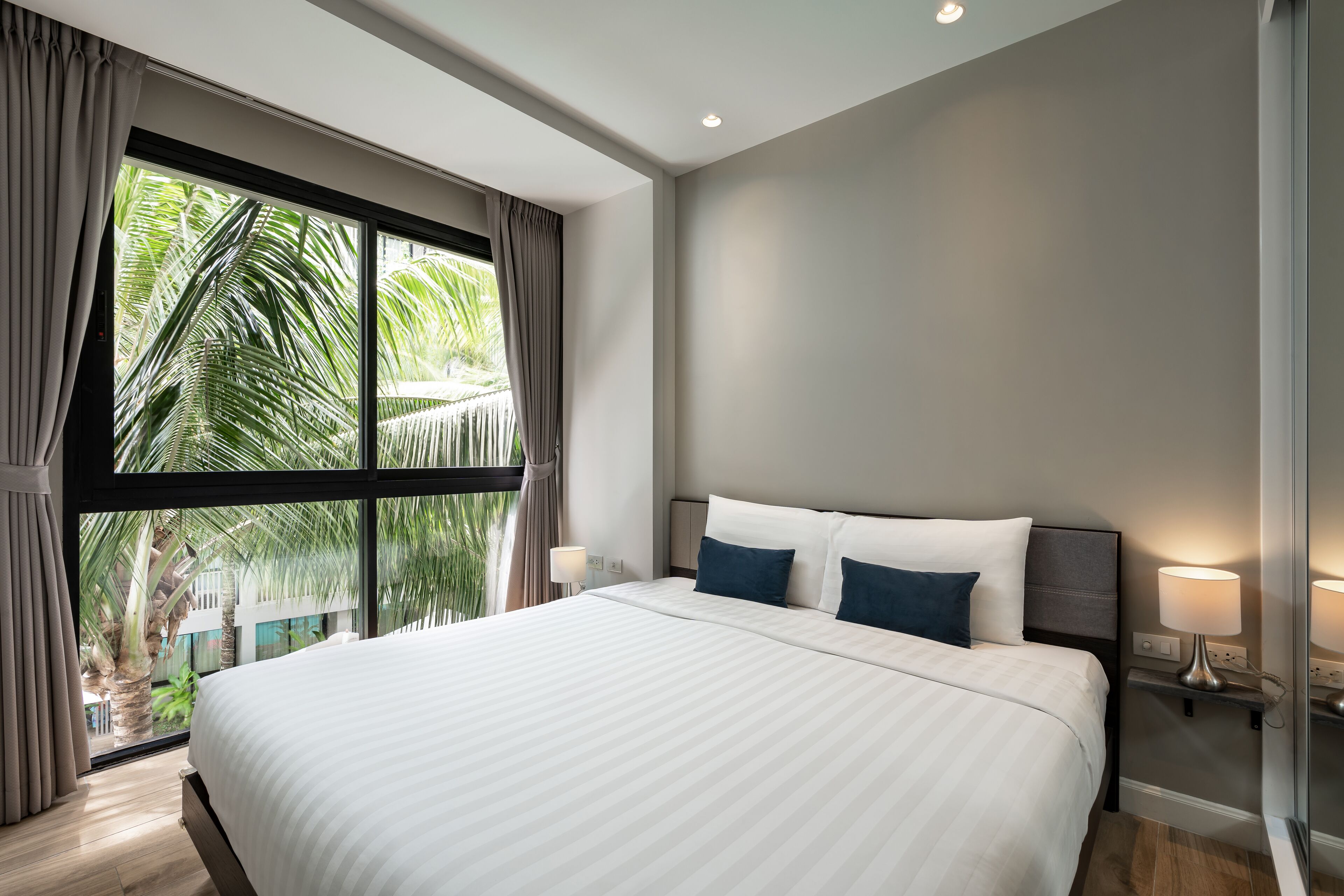 A modern hotel room with a large bed, two bedside lamps, and a view of palm trees through a large window.
