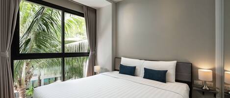 Ein modernes Hotelzimmer mit einem großen Bett, zwei Nachttischlampen und Blick auf Palmen durch ein großes Fenster.
