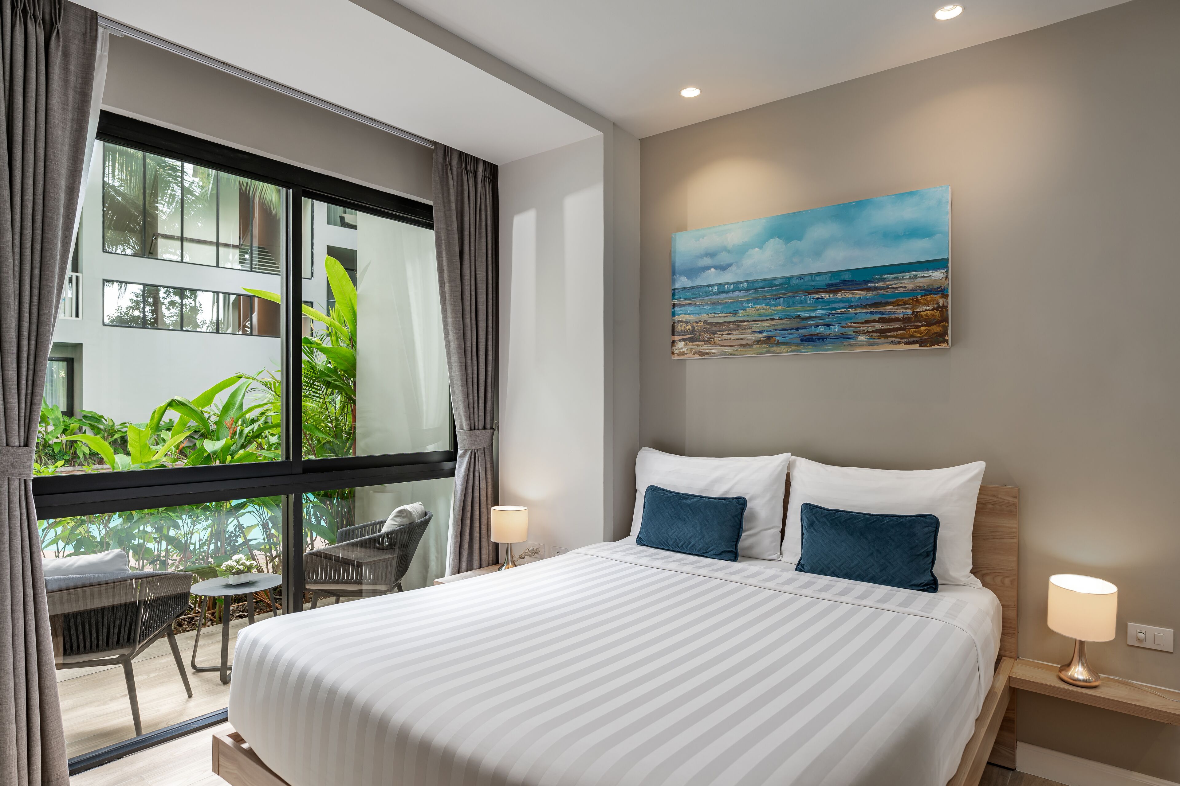 Una habitación de hotel moderna con una cama grande, dos almohadas, una mesita de noche con una lámpara, un cuadro de una playa y vistas al verdor a través de un ventanal amplio.