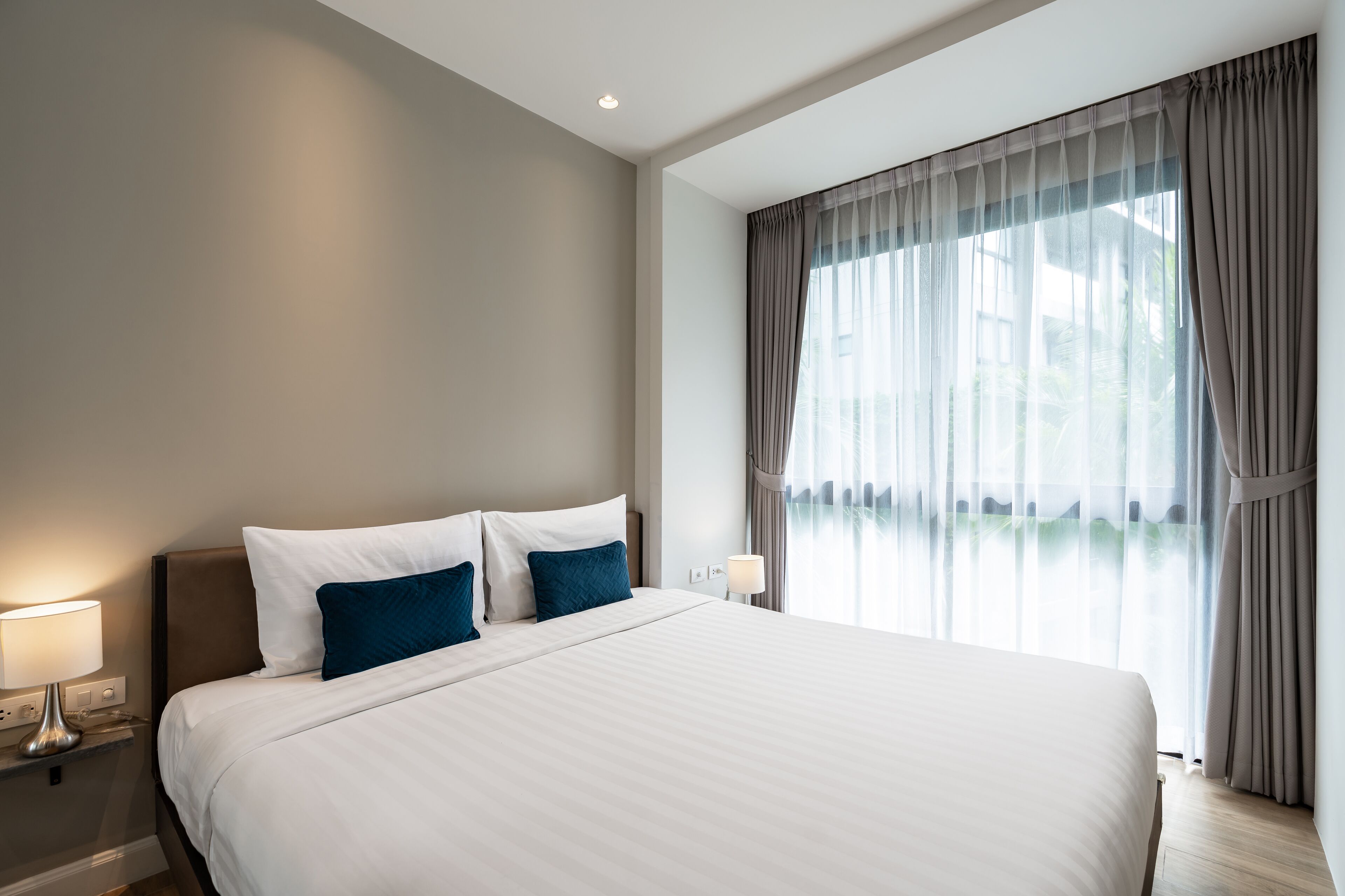 A hotel room with a large bed, a nightstand with a lamp, and a window with sheer curtains.