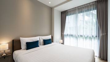 A hotel room with a large bed, a nightstand with a lamp, and a window with sheer curtains.