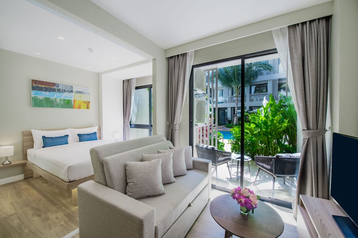Quarto de hotel moderno com cama, sofá, pequena mesa com flores e varanda com vista para a piscina.