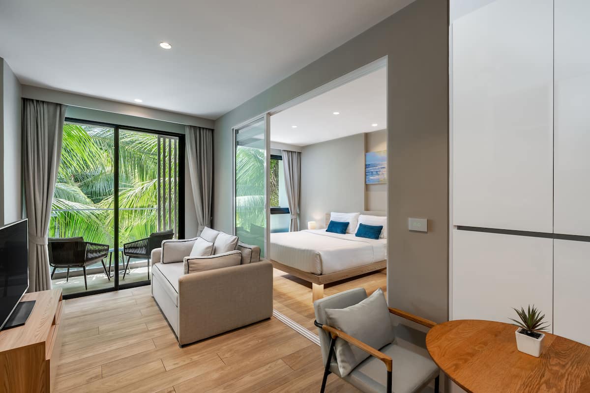 Quarto de hotel moderno com sofá, cama, TV, mesa de jantar e varanda com vista para o verde.
