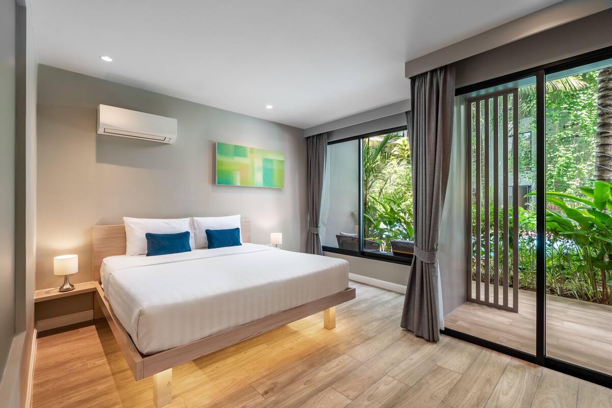 Quarto de hotel moderno com uma cama grande, mesa de cabeceira, ar condicionado fixado na parede, janela com vista para vegetação e um quadro na parede.