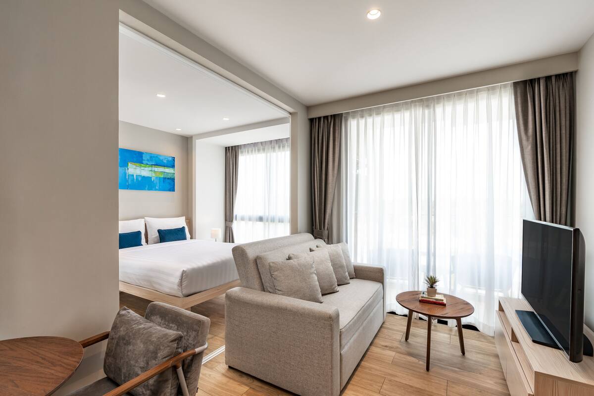 Quarto de hotel moderno com sofá, televisão, mesinha e cama com uma janela ampla.