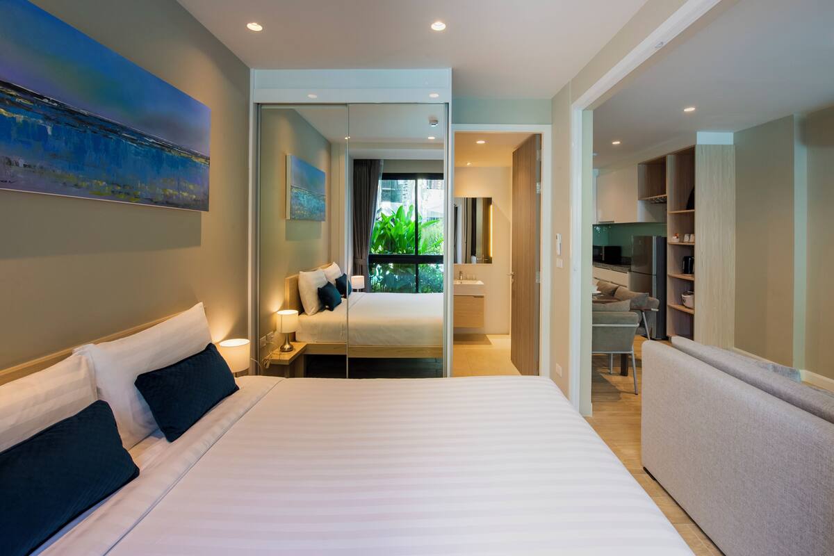 Quarto de hotel moderno com cama grande, mesa de cabeceira, espelho, janela com vista para o verde e área de cozinha compacta.