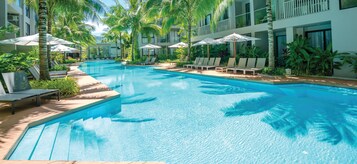 Piscina com espreguiçadeiras e guarda-sóis, cercada por palmeiras e um edifício moderno.