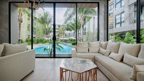 A modern living room with a pool view, featuring a sofa set, a marble coffee table, and large windows overlooking a pool area with palm trees and a building in the background. - Diamond Resort Phuket Official Account (Choeng Thale)