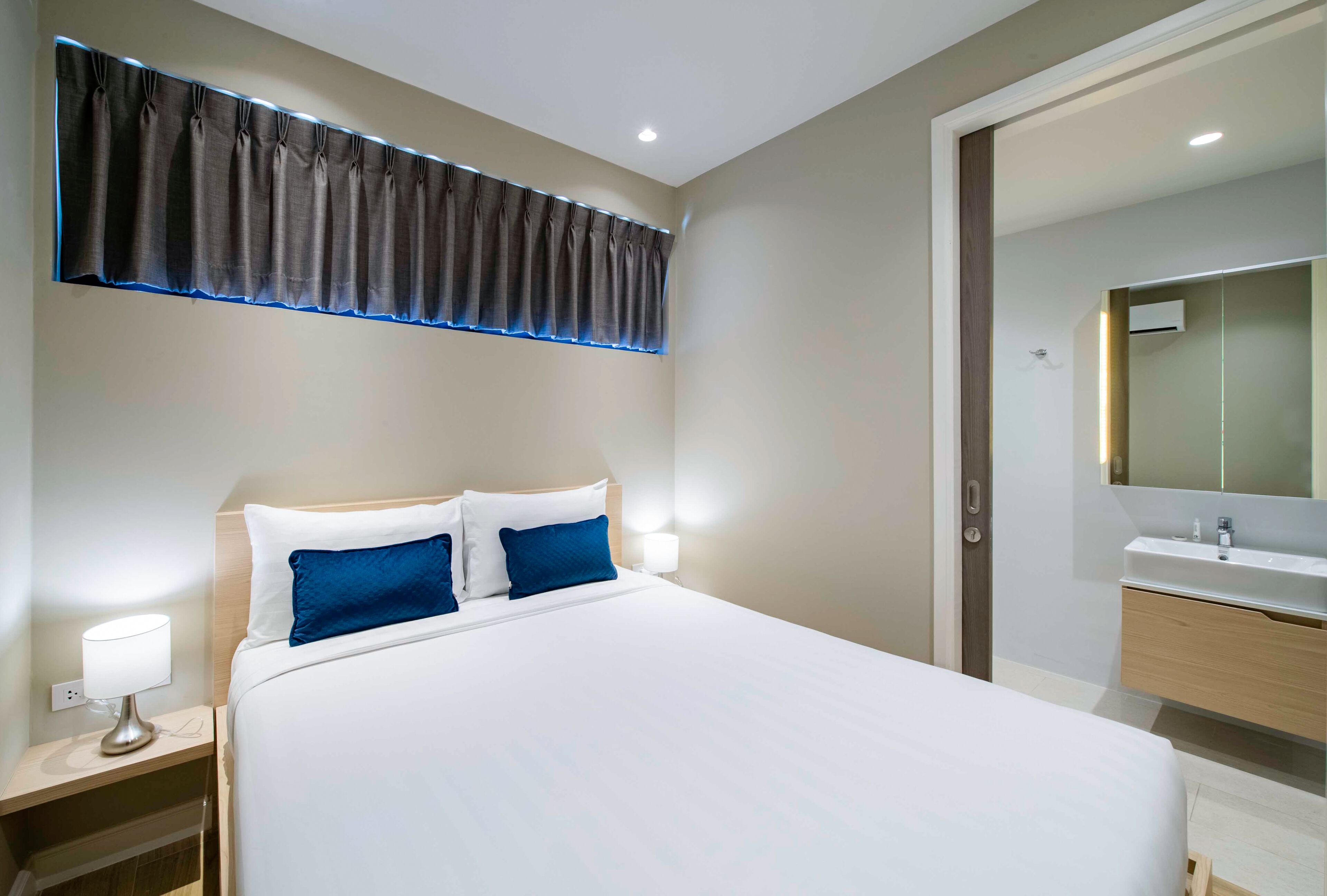 A modern hotel room with a large bed, bedside lamps, a nightstand, a bathroom with a mirror and sink, and a window with curtains.