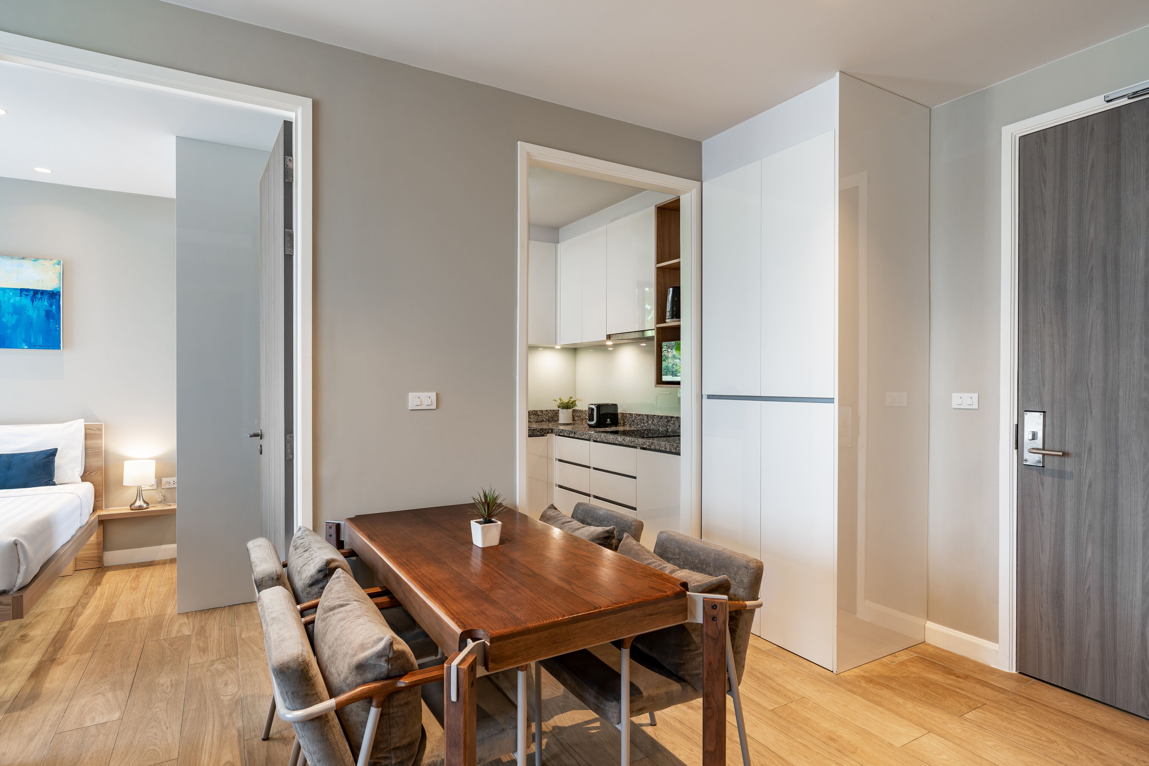 A modern apartment with a wooden dining table, cushioned chairs, a small kitchen area, and a separate living space with a bed and a nightstand.
