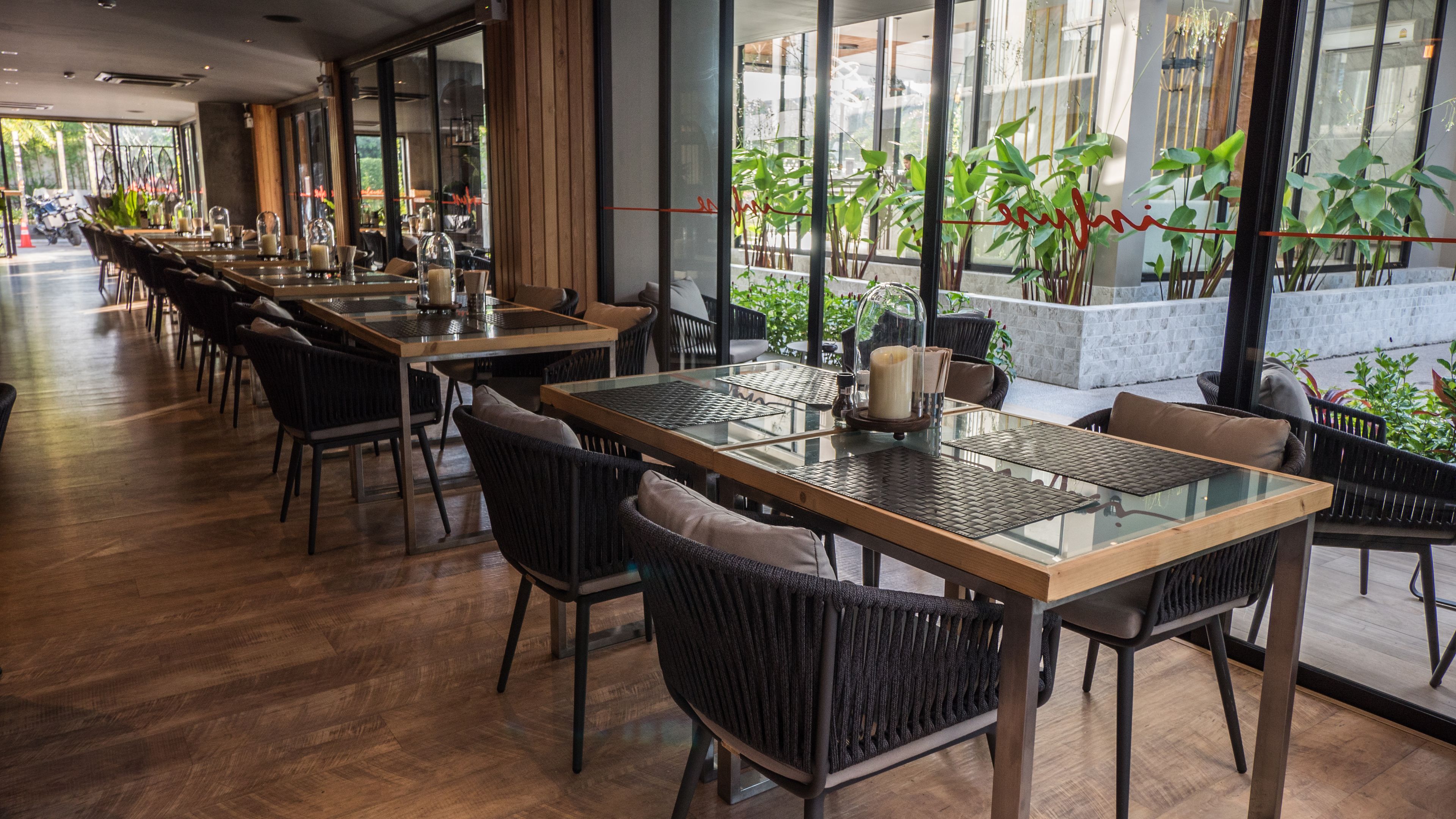 A restaurant with long tables and chairs, large windows, and indoor plants.