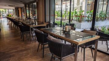 A restaurant with long tables and chairs, large windows, and indoor plants.