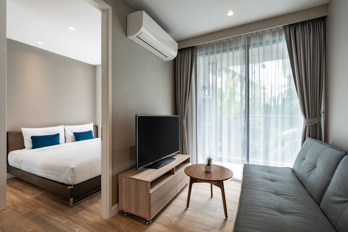 Quarto de hotel moderno com cama, TV sobre suporte de madeira, sofá e janela com cortinas.