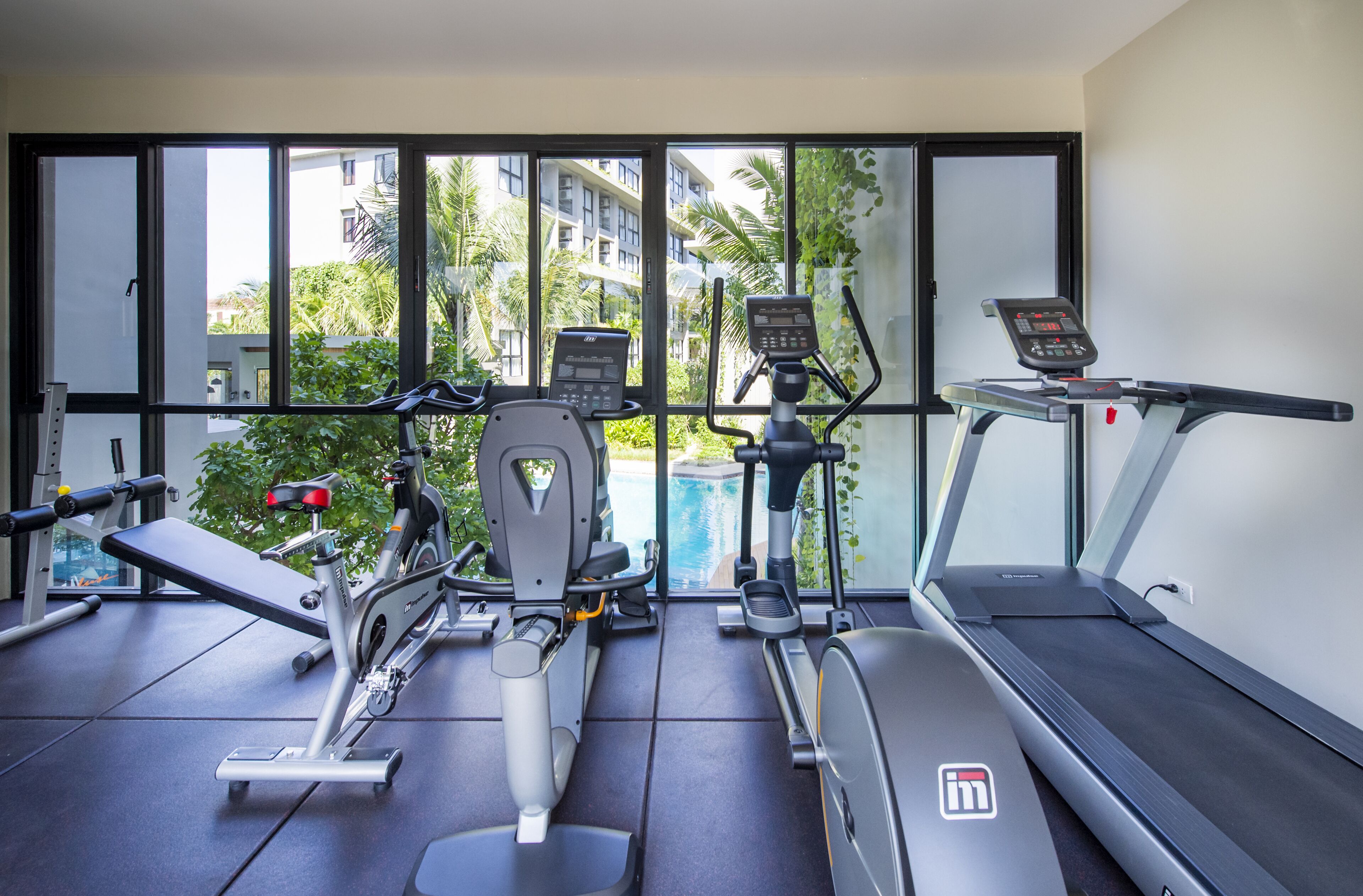 A gym with cardio machines, a bench, and a treadmill, with large windows overlooking a pool and greenery.