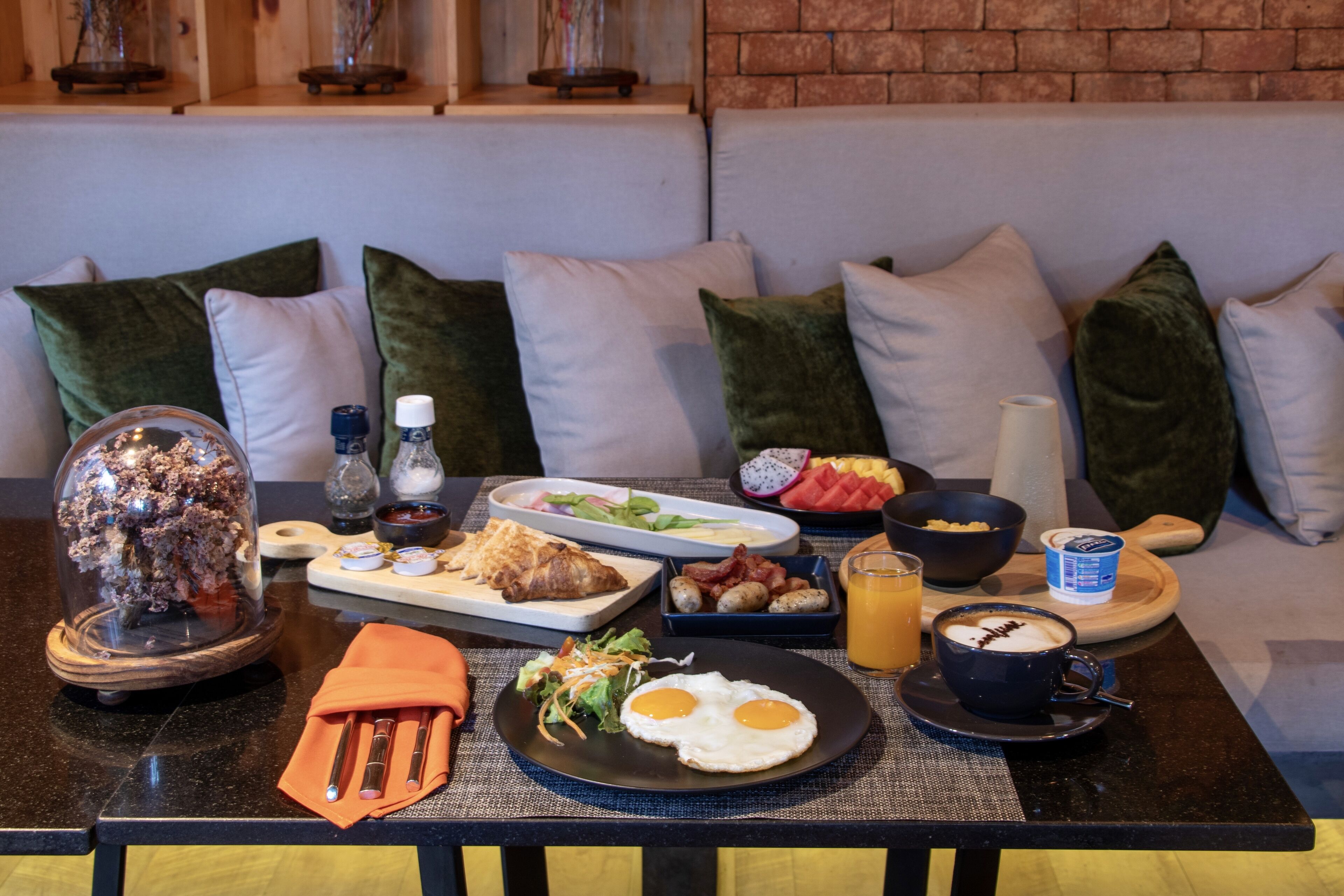 A breakfast setting with a variety of food items on a table, including eggs, bacon, toast, and fruit, in a room with a brick wall and green pillows.