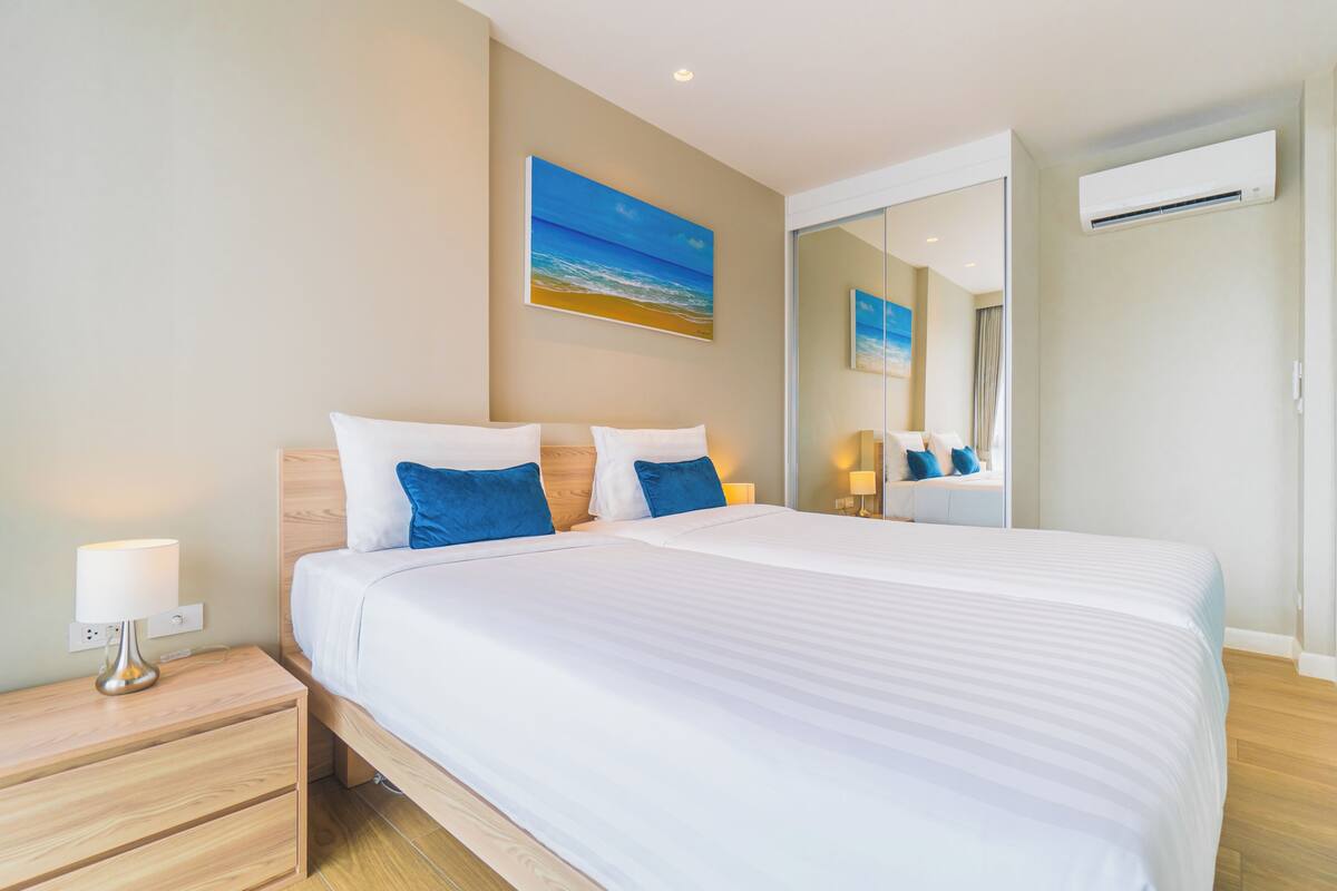 Quarto de hotel com cama, mesa de cabeceira, luminária e uma pintura de uma praia.