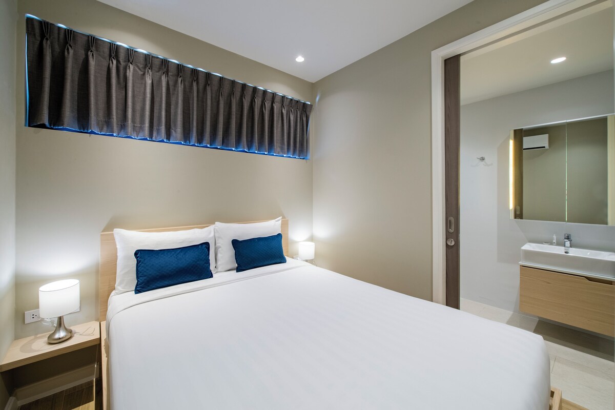 Habitación de hotel con una cama grande, lámparas de noche, una mesita de noche y un baño con espejo y lavamanos.