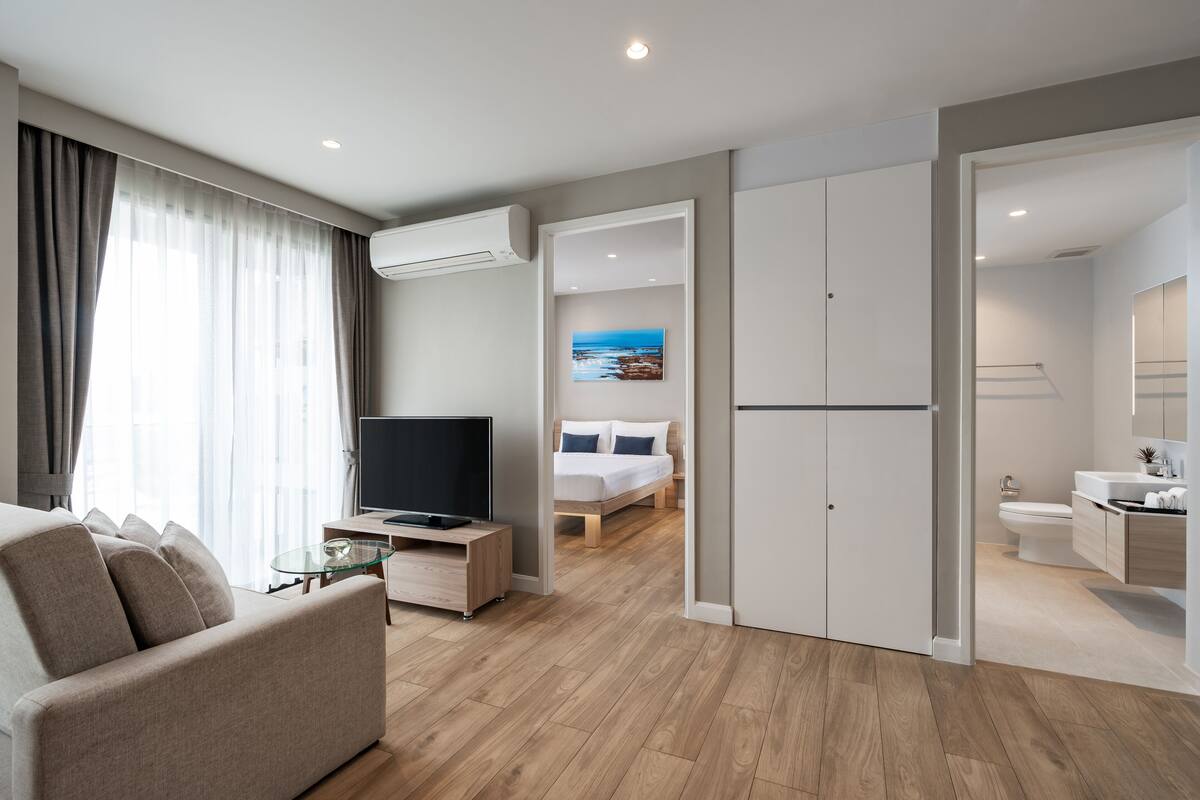 Quarto de hotel moderno com sofá, TV, piso de madeira, cama com pintura e banheiro com pia e vaso sanitário.