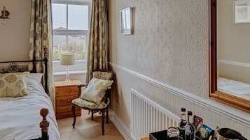 Quarto com cama, cadeira, mesa de cabeceira, radiador e janela com cortinas.