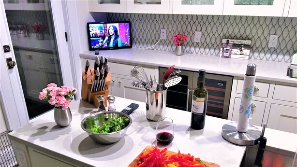 A modern kitchen with a white countertop, a bowl of greens, a cutting board with sliced vegetables, a wine bottle, and a TV displaying a show.