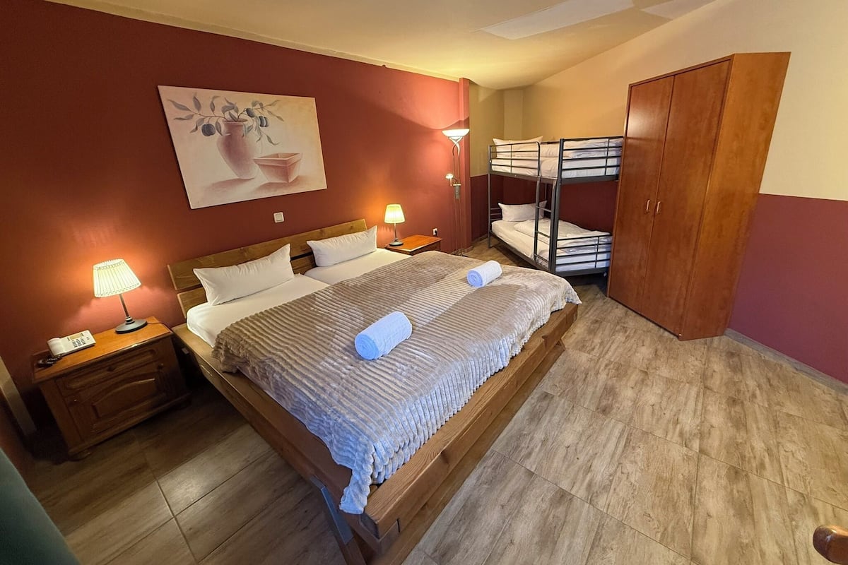 A hotel room with a wooden bed, two bedside tables with lamps, a bunk bed, and a wooden wardrobe.