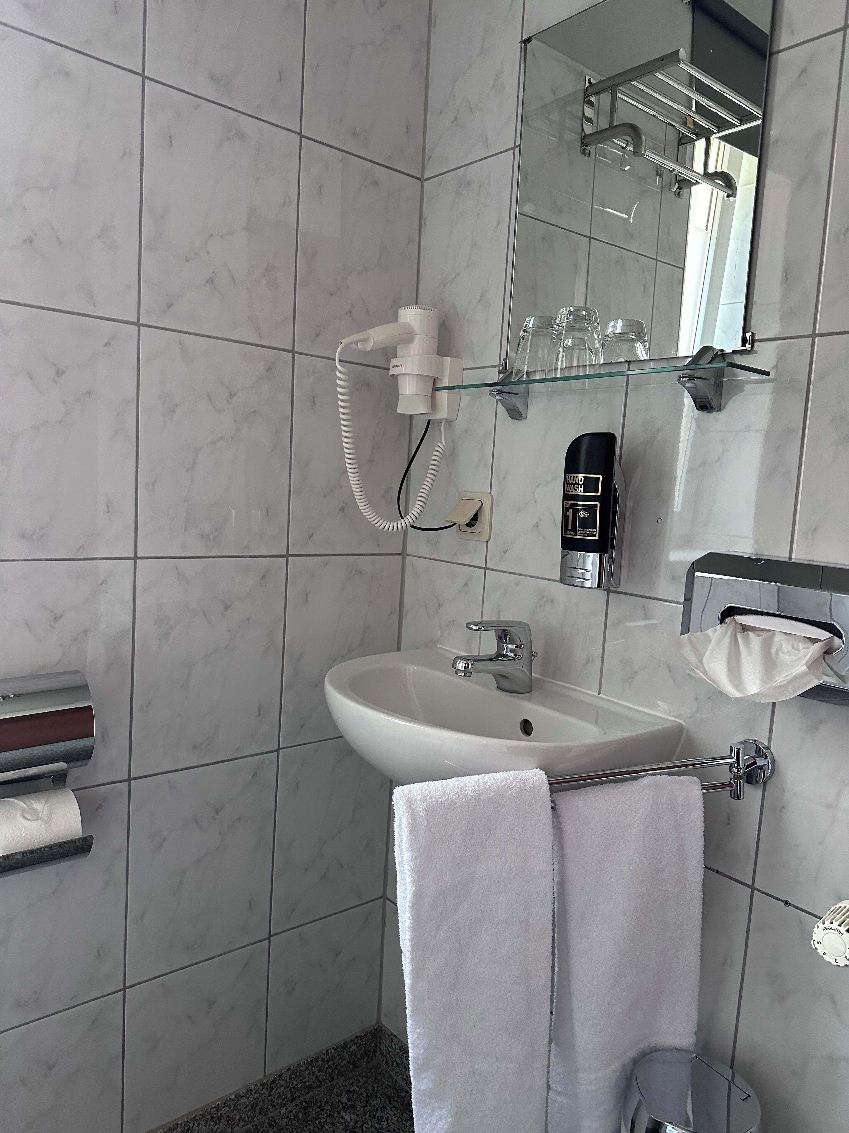 Ein modernes Badezimmer mit einem weißen Waschbecken, einem Handtrockner, einer Duschkabine aus Glas und einem Handtuchhalter.