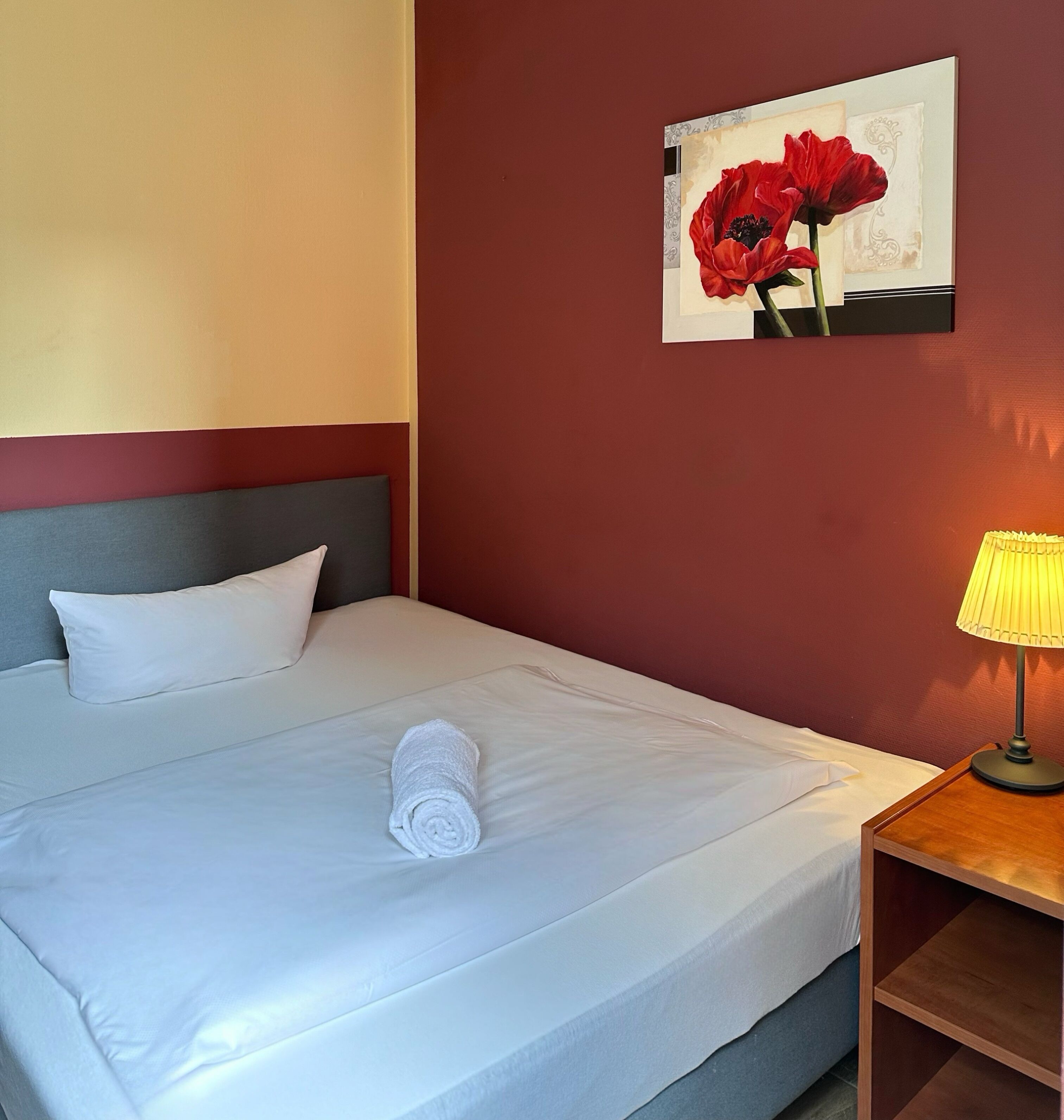Ein ordentlich gemachtes Bett mit weißen Leinen, ein zusammengerolltes Handtuch und ein einzelnes weißes Kissen. Über dem Bett hängt ein gerahmtes Bild von roten Blumen.