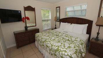 A bedroom with a large bed, a TV mounted on the wall, a dresser, and a bedside table with a lamp.