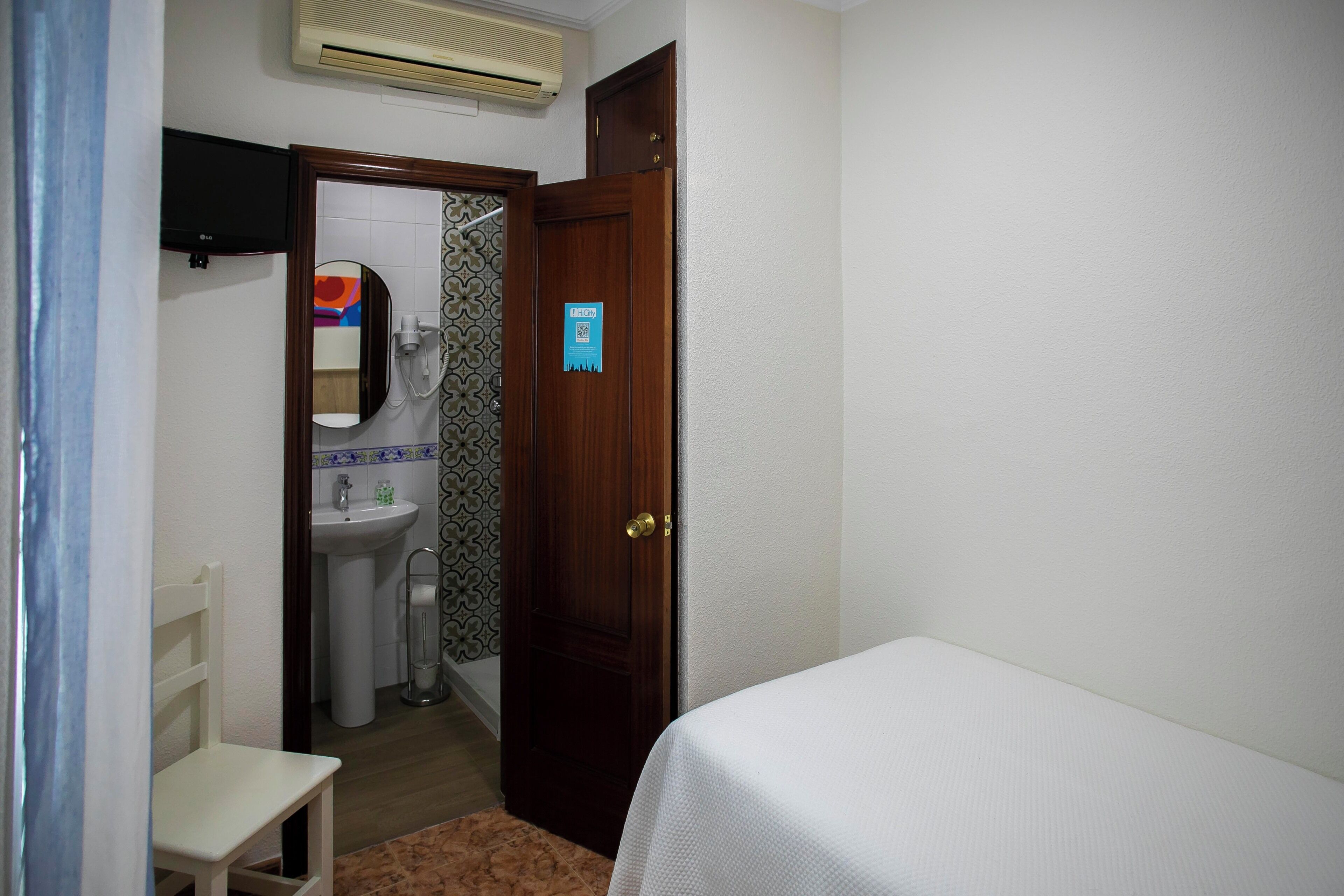 A bedroom with a bed, a wooden door, a bathroom with a sink and mirror, and a TV mounted on the wall.