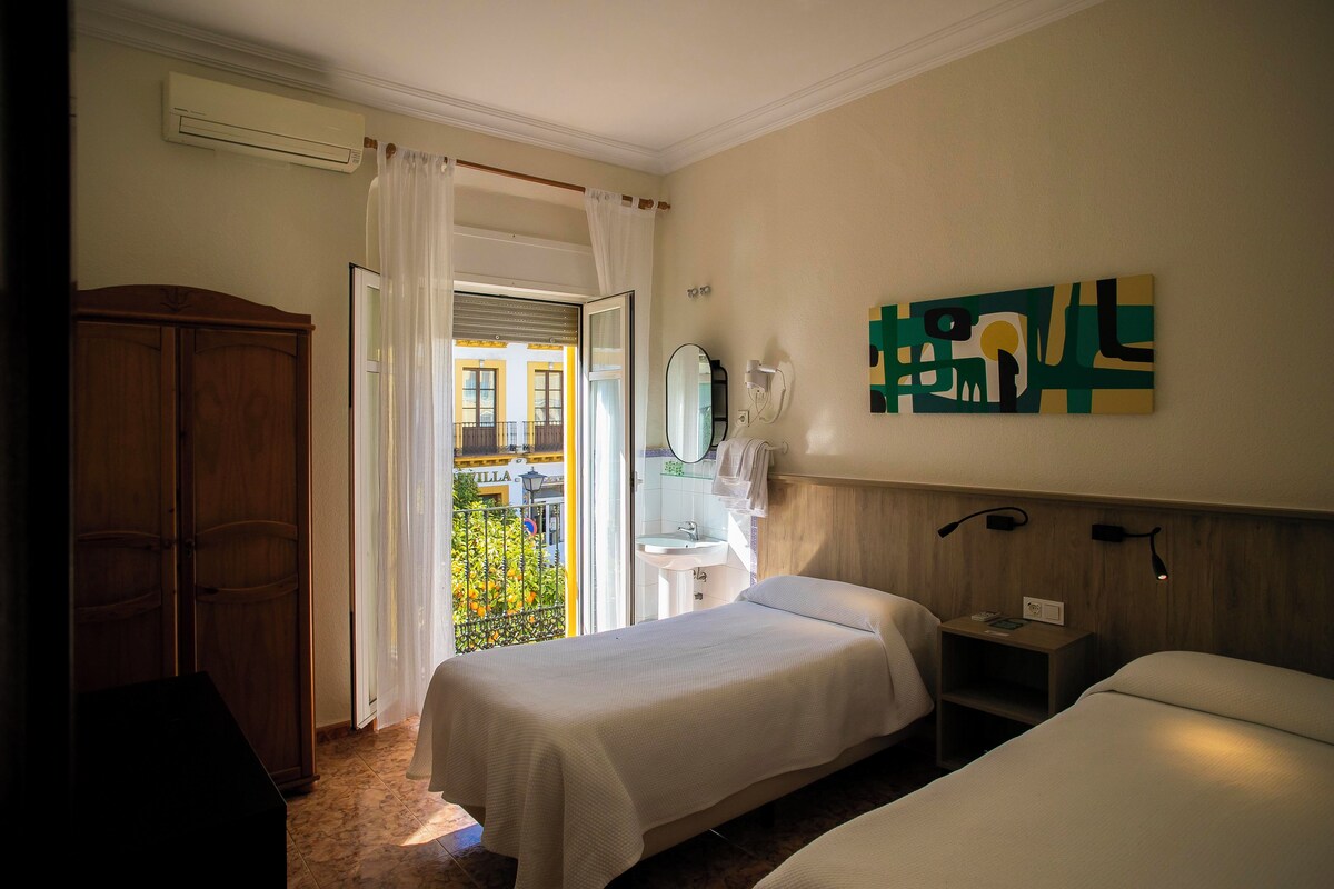 A hotel room with two beds, a wooden wardrobe, a bathroom with a sink and mirror, and a balcony with a view of buildings.