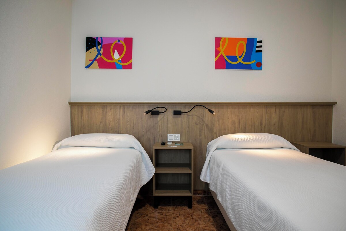 A hotel room with two single beds, a wooden headboard, a nightstand with a lamp, and two abstract paintings on the wall.