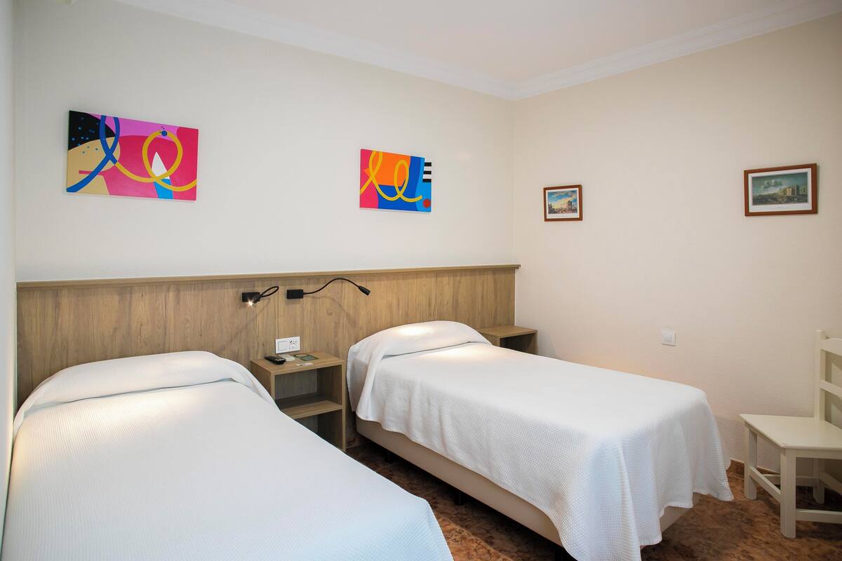 A hotel room with two single beds, a wooden headboard, a nightstand with a lamp, and two wall-mounted artworks.