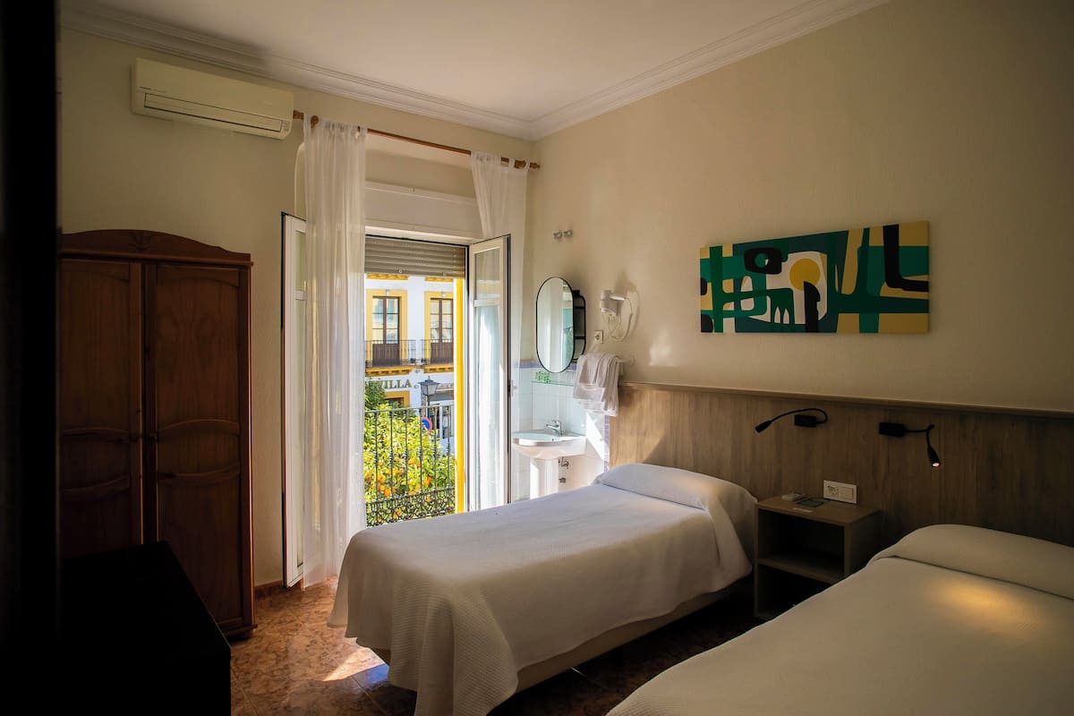 A hotel room with two beds, a wooden wardrobe, a bathroom with a sink and mirror, and a balcony with a view of buildings.