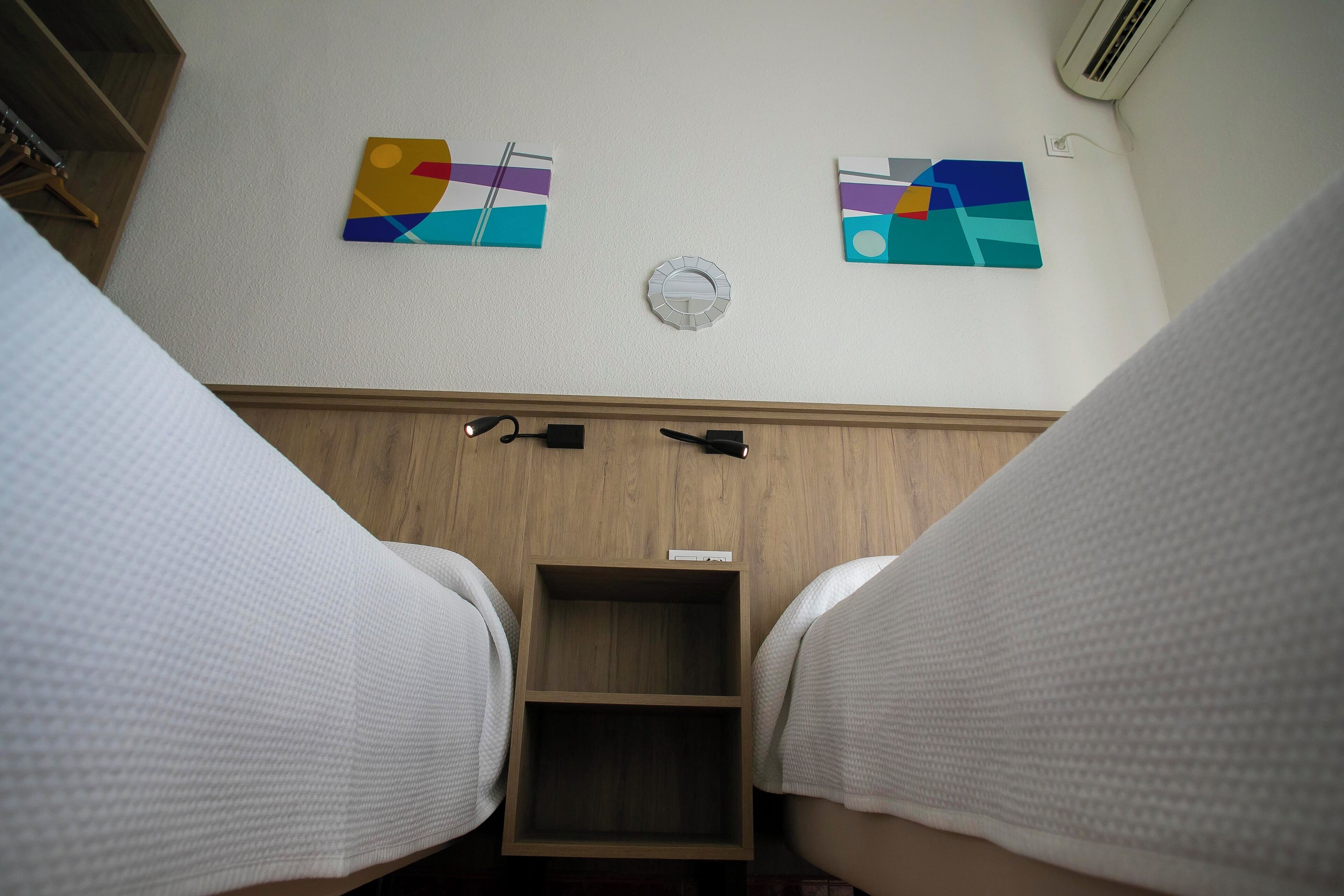 A hotel room with two beds, wooden headboards, a small wooden shelf, and two abstract wall art pieces.