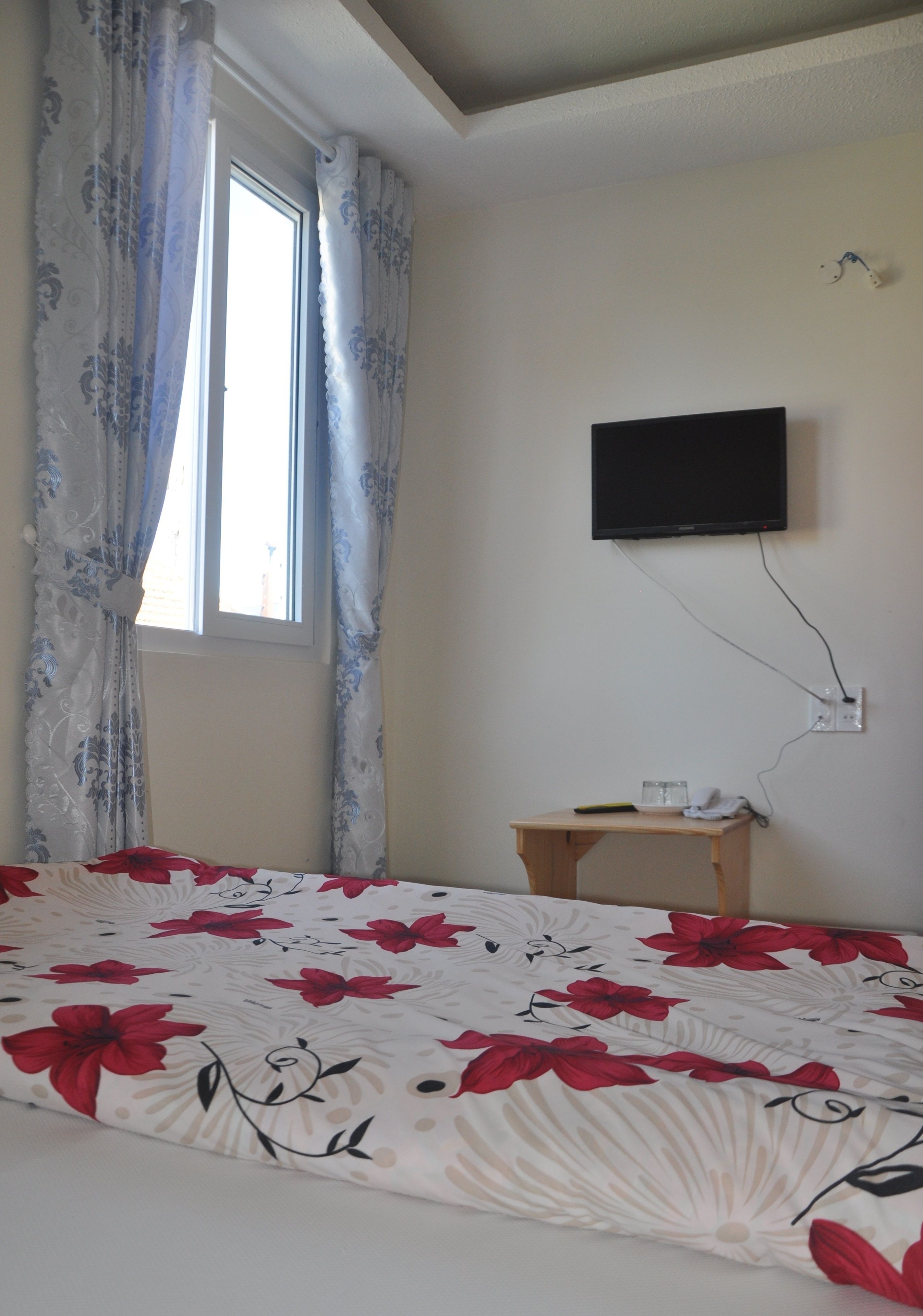 Ein Schlafzimmer mit einem Bett, einem Fenster mit Vorhängen, einem an der Wand montierten Fernseher und einem kleinen Holztisch mit Fernbedienung und Gläsern.