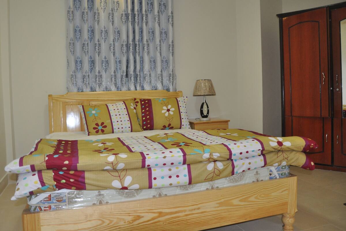 A neatly made bed with a floral bedspread, a wooden headboard, a bedside lamp, and a wardrobe in a room with patterned curtains.