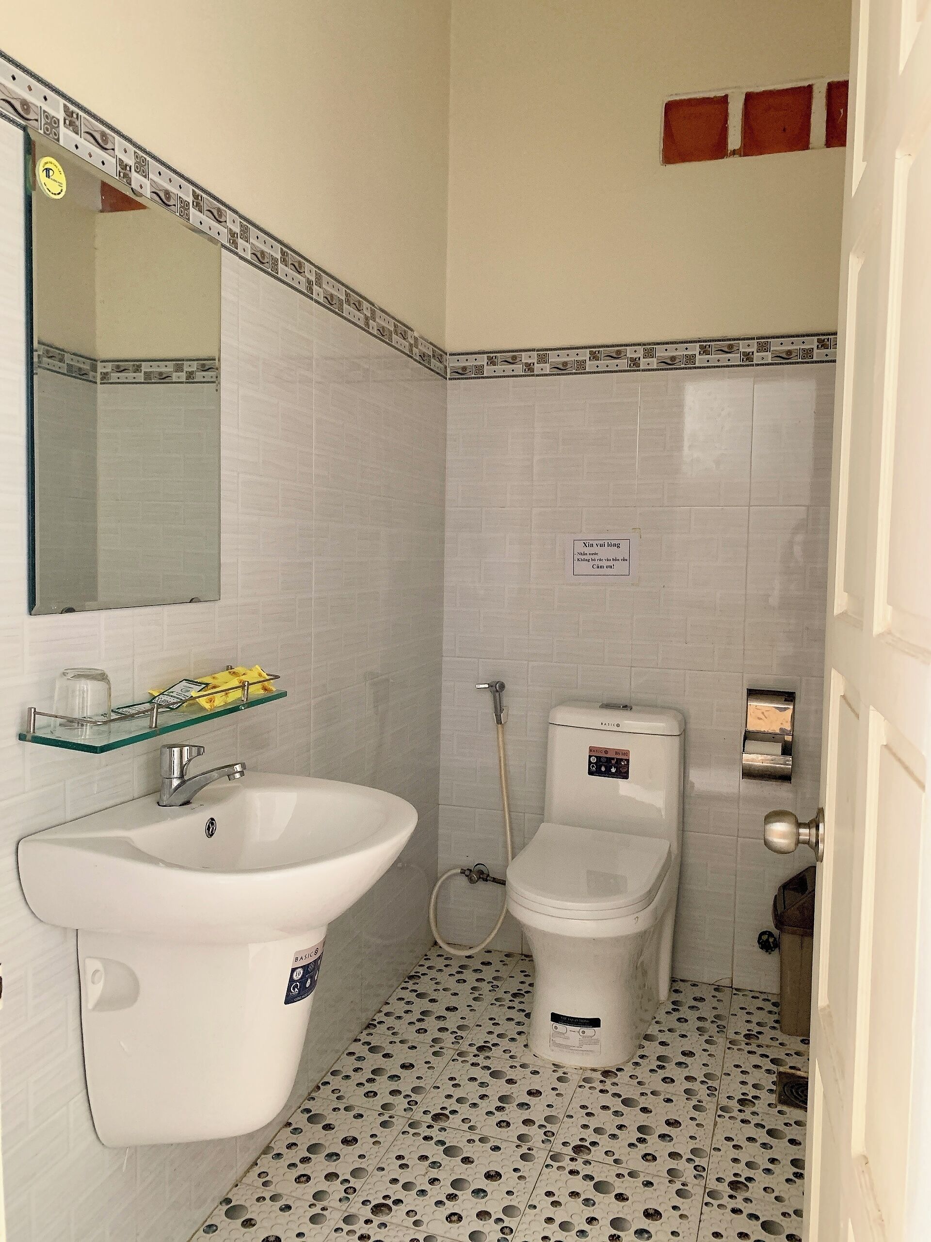 A bathroom with a white sink, a toilet, and a shower. There is a mirror above the sink and a sign on the wall.