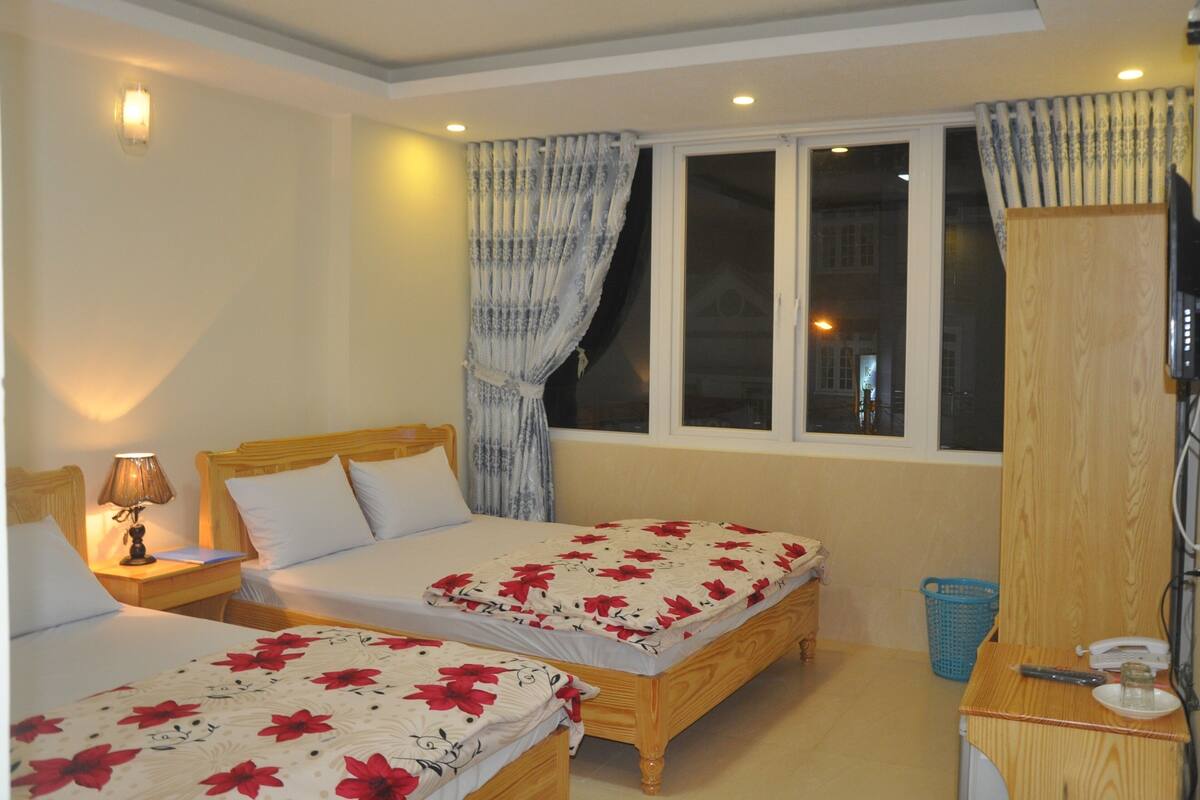 A bedroom with two beds, a desk, a lamp, a TV, and a window with curtains.